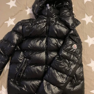  Moncler Maya - Säljer en snygg svart dunjacka från Moncler i mycket bra skick. Jackan har en glansig finish och är perfekt för kalla vinterdagar. Den har en dragkedja framtill och två praktiska fickor. Moncler-loggan syns tydligt på ärmen. Perfekt för dig som vill hålla stilen i vinter! 🖤