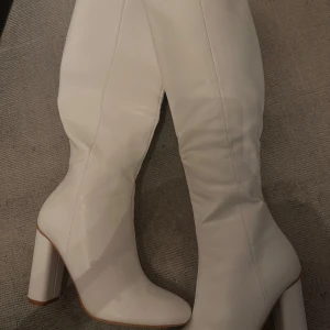 Vita stövlar med klack, vita boots - EJ ANVÄND!  Storlek 3 uk, 35-36 eu. Säljer ett par snygga vita stövlar i syntetmaterial. De har en hög klack och når till knäna, vilket ger en elegant look. Perfekta för både fest och vardag! De är i nytt skick och har en dragkedja på insidan för enkel på- och avtagning. Skorna kommer i orginalparketet med kartong och plastet i så de inte viks eller förstörs. 
