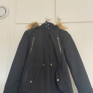 Svart parkajacka med fuskpäls - Säljer en svart parkajacka med fuskpäls på luvan. Jackan har dragkedja och knappar framtill samt praktiska fickor med knappar. Insidan är fodrad med mjukt material för extra värme. Perfekt för kalla vinterdagar! 🖤