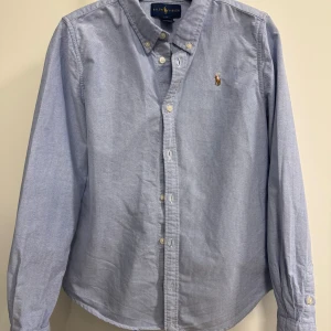 Blå skjorta från Ralph Lauren till flicka - Jättefin ljusblå skjorta i något insvängd modell. Använd 2-3 ggr. Strl 14 år men den är liten och passar 146/152 bra. Mycket fint skick, inköpt på NK Kids för ett par år sedan.