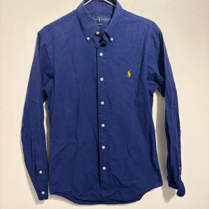 Ralph Lauren skjorta - Säljer en snygg blå skjorta från Ralph Lauren i slim fit. Skjortan har långa ärmar och klassiska knappar framtill. Den är dekorerad med det ikoniska gula broderade logotypen på bröstet. Perfekt för både vardag och fest!                                  299kr ny pris 1299