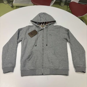 Grå Burberry hoodie med dragkedja - Säljer en snygg grå hoodie från Burberry med dragkedja och huva. Insidan av huvan har det klassiska Burberry-mönstret. Perfekt för en avslappnad stil och passar till alla årstider. Den är i nytt skick och har en normal passform.