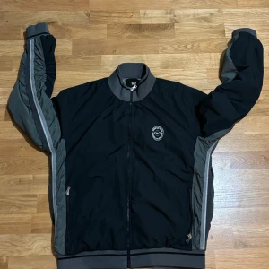 Svart vindjacka från Dolce & Gabbana - Säljer en snygg svart bomberjacka från Dolce & Gabbana i storlek 58/L jackan är en väldigt gammal modell tror jag har ingen aning. En av dragkedjor fickorna går inte att stänga.  Jackan har grå detaljer och en dragkedja framtill. Den är perfekt för höst och vår med sitt stilrena utseende och bekväma passform. Insidan har ett coolt tryck med text. Passar både till vardags och fest!