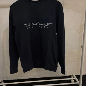 Mörkblå Hugo boss sweat shirt - Säljer en stilren svart tröja från Hugo Boss. Den har en klassisk design med långärmade ärmar och en rund halsringning. Perfekt för både vardag och lite finare tillfällen. Hugo Boss-loggan är broderad på framsidan. Passar bra till jeans eller chinos.