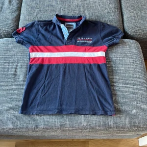 Mörkblå pikétröja från Race Marine - Säljer en snygg mörkblå pikétröja från Race Marine i storlek XS. Den har röda och vita ränder över bröstet och broderad text på framsidan. Tröjan är kortärmad och perfekt för en avslappnad stil. Passar både till vardags och lite finare tillfällen. 🏄‍♂️Pris kan alltid förhandlas!