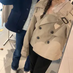 Säljer en snygg beige trenchcoat med bälte och dubbelknäppning. Jackan har en klassisk stil med en modern twist och är perfekt för höst och vår. Den är i mycket bra skick och passar till både vardag och fest. Perfekt för dig som vill ha en stilren look! Pris kan diskuteras!!