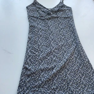 Blommig klänning med spetsdetaljer - Säljer en superfin blommig klänning med smala axelband och spetsdetaljer längs nederkanten. Klänningen har en omlott-design upptill och är perfekt för sommardagar. Materialet känns mjukt och bekvämt, och den har en svart och vit färgkombination som är lätt att matcha med andra plagg.