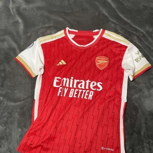 Röd Arsenal fotbollströja - Säljer en snygg röd Arsenal fotbollströja från Adidas. Den har korta vita ärmar med gulddetaljer och klubbens emblem på bröstet. Tröjan är i mycket bra skick och har ett coolt mönster. Perfekt för alla Arsenal-fans! ⚽️