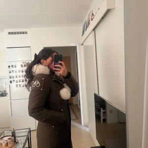 Grön parkajacka med pälskrage - Säljer en snygg olive grön parkajacka från Moose Knuckles i mycket bra skick. Jackan har en fluffig vit pälskrage och matchande detaljer på huvan. Perfekt för kalla vinterdagar med sin varma och stiliga design. Den har en dragkedja och knappar framtill för extra skydd mot kylan. Pris går att diskuteras vid snabb köp! 