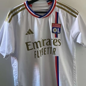 Olympique Lyonnais fotbollströja - Säljer en vit Olympique Lyonnais fotbollströja från Adidas i ny skick. Tröjan är äkta och aldrig använd pga för stor! Tröjan har korta ärmar och guldiga detaljer med klubbens emblem på bröstet. Den har också ränder i rött och blått runt kragen och längs sidan. Perfekt för fans av laget eller samlare! Bara att höra av sig vid minsta fundering! Storlek S, passar fint till M