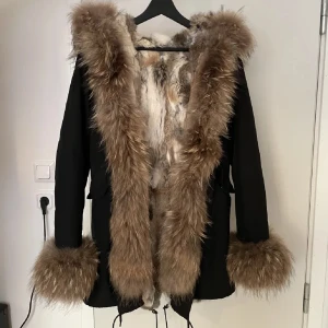 Svart jacka med pälsdetaljer - Säljer en supermysig svart jacka med fluffiga pälsdetaljer runt huva, ärmslut och insida. Perfekt för kalla vinterdagar när du vill hålla dig varm och stilren. Jackan har en normal passform och är i mycket bra skick. Pälsen ger en lyxig känsla och är i olika naturliga nyanser.