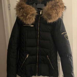 Svart dunjacka med pälskrage från Rockandblue - Säljer en snygg svart dunjacka från Rockandblue i mycket bra skick. Jackan har en fluffig pälskrage i äkta rävpäls och guldfärgade dragkedjor. Perfekt för kalla vinterdagar! 🖤