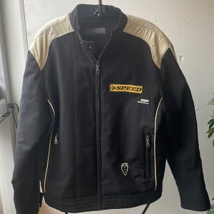 Svart jacka från Jack & Jones - Säljer en cool svart jacka från Jack & Jones med beige läder detaljer och texten 'SPEED' på bröstet. Jackan har dragkedja och två fickor med dragkedjor. Perfekt för höst och vår, med en sportig look. Passar bra för både vardag och fest!