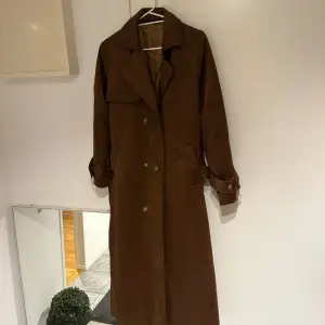 Säljer en snygg brun trenchcoat, oanvänd . Den har långa ärmar och knappar framtill samt ett bälte i midjan för en klassisk look. Perfekt för höst och vår när du vill vara både stilren och varm. 