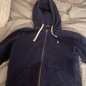 Mörkblå huvtröja från Polo Ralph Lauren - Säljer en snygg mörkblå huvtröja från Polo Ralph Lauren i storlek M passar perfekt som S. Tröjan har en dragkedja framtill och en liten gul logga på bröstet. Perfekt för höst och vår, med långa ärmar och en skön huva med vita snören. Passar både till vardags och lite mer avslappnade tillfällen.