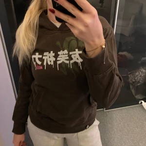 Brun hoodie med tryck - Säljer en brun hoodie med coolt tryck i vitt och grönt. Den har en känguruficka och är perfekt för en avslappnad stil. Passar bra till både vardag och chillkvällar. Grymt skön och i bra skick!
