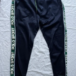 Mörkblå träningsbyxor från Polo Sport - Säljer ett par mörkblå träningsbyxor från Polo Sport med gröna och vita detaljer längs sidorna. Byxorna har en normal passform och är perfekta för träning. De är i mycket bra skick och har en elastisk midja med snörning för bästa komfort.