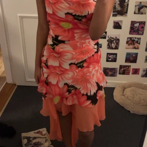 Blommig klänning med volangdetaljer - Säljer en färgglad blommig klänning med stora orange och rosa blommor. Klänningen har volangdetaljer och är ärmlös, perfekt för sommarens fester. Den har en figurnära passform och är i mycket bra skick.