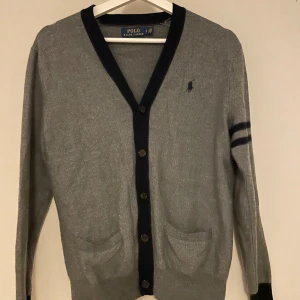 Grå kofta från Polo Ralph Lauren - Säljer en stilren grå kofta från Polo Ralph Lauren. Den har långa ärmar och en klassisk v-ringning med knappar framtill. Två fickor fram och en diskret broderad logga på bröstet. Perfekt för både höst och vår!