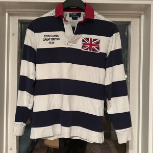 Randig pikétröja från Polo Ralph Lauren - Säljer en snygg randig pikétröja från Polo Ralph Lauren i marinblått och vitt. Tröjan har en röd krage och broderad Union Jack-flagga på bröstet.  Den är i bra skick, då jag inte har använt den så mycket.