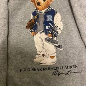 Säljer en superfin grå hoodie från Ralph Lauren med det ikoniska Polo Bear-trycket på framsidan. Perfekt för en avslappnad stil med en touch av lyx. Hoodien har långa ärmar och en bekväm passform. Perfekt för höst och vinter!