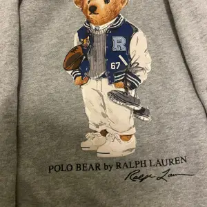 Säljer en superfin grå hoodie från Ralph Lauren med det ikoniska Polo Bear-trycket på framsidan. Perfekt för en avslappnad stil med en touch av lyx. Hoodien har långa ärmar och en bekväm passform. Perfekt för höst och vinter!