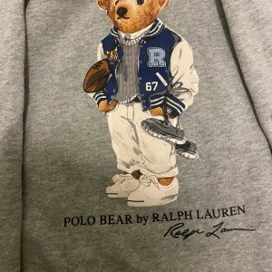 Grå hoodie med Polo Bear från Ralph Lauren - Säljer en superfin grå hoodie från Ralph Lauren med det ikoniska Polo Bear-trycket på framsidan. Perfekt för en avslappnad stil med en touch av lyx. Hoodien har långa ärmar och en bekväm passform. Perfekt för höst och vinter!