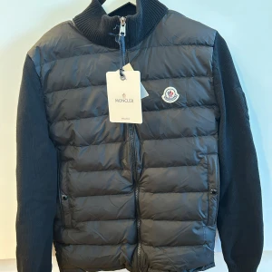 Svart Moncler jacka - Säljer en snygg svart Moncler jacka i mycket bra skick. Storlek M men sitter som S.    Den har en quiltad framsida med dragkedja och stickade ärmar. Perfekt för höst och vinter! Jackan har en klassisk Moncler-logga på bröstet och en ribbad krage för extra stilpoäng.