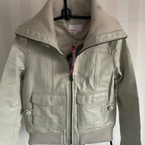 Beige skinnjacka från Zoul - Snygg beige skinnjacka från Zoul i storlek 36. Jackan har en cool dragkedja och ribbade detaljer vid krage, ärmslut och nederkant. Den har praktiska fickor med knappar framtill. Perfekt för höst och vår!