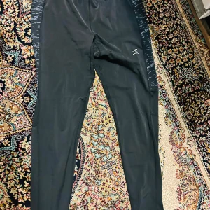 Svarta träningsbyxor från Energetics - Säljer ett par svarta träningsbyxor från Energetics i storlek XL. Byxorna har en tight passform och är gjorda av ett syntetmaterial med DRYPLUSecco-teknologi för att hålla dig torr under träning. De har snygga mönstrade paneler längs sidorna och dragkedjor vid bensluten för extra ventilation.