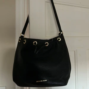 Svart axelväska från Michael Kors - Svart axelväska från Michael Kors i skinn. Väskan har guldfärgade detaljer och en praktisk dragkedja inuti. Perfekt för både vardag och fest! 