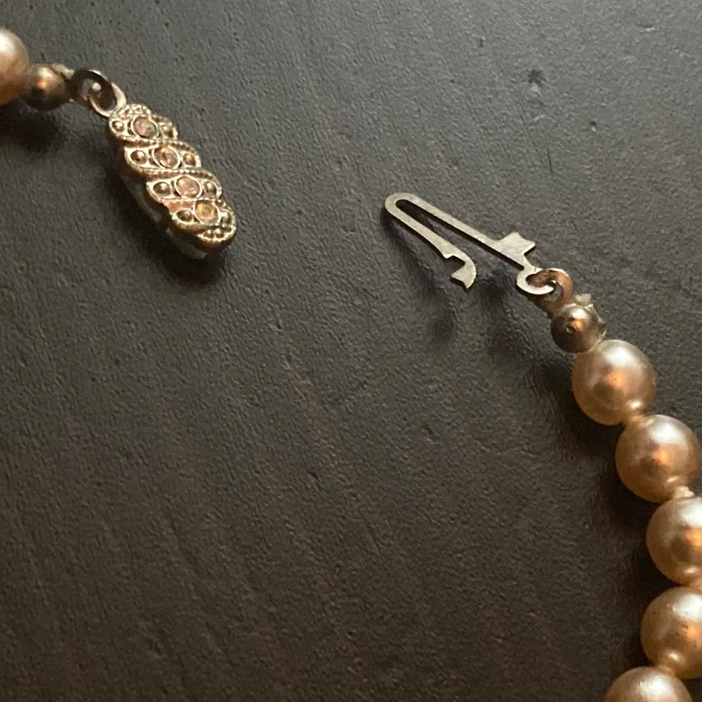 Äldre halsband med tyngre ljusbeiga plastpärlor och silverfärgat lås med strass. Finns slitage på pärlorna och låset har missfärgningar. Längd: 40 cm, bredd: 6 mm.. Asusteet.