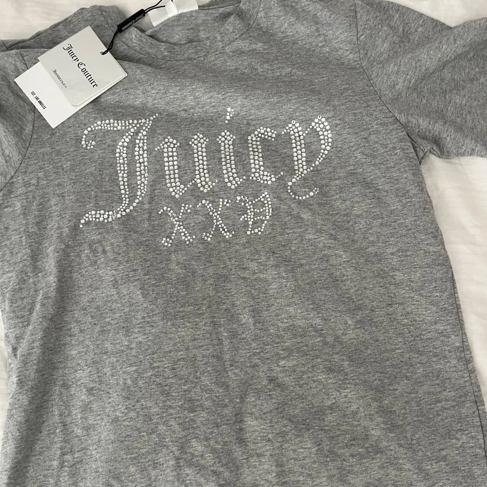 Snygg grå t-shirt från Juicy Couture i storlek S. Den är gjord av mjuk bomull och har ett glittrigt Juicy-märke på framsidan. Perfekt för en avslappnad men ändå stilren look. Passar bra till både jeans och kjol!. T-shirts.