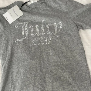 Grå t-shirt från Juicy Couture - Snygg grå t-shirt från Juicy Couture i storlek S. Den är gjord av mjuk bomull och har ett glittrigt Juicy-märke på framsidan. Perfekt för en avslappnad men ändå stilren look. Passar bra till både jeans och kjol!