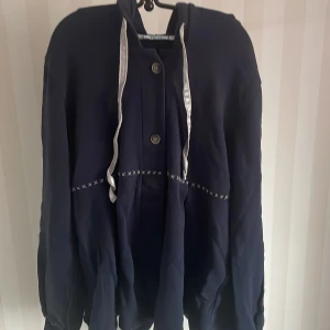 Mörkblå jacka från Disney Signature - Säljer en mörkblå jacka från Disney Signature i storlek 44. Jackan har långa ärmar och knappar framtill. Den är dekorerad med ett diskret broderi och har en knytning i midjan. Perfekt för höst och vår!