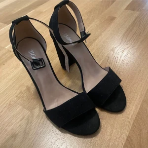 Svarta klackskor med öppen tå - Snygga svarta klackskor med öppen tå och justerbart spänne runt ankeln. Perfekta för festliga tillfällen! De har en stilren design med en bred klack som ger bra stabilitet. Materialet ser ut att vara syntetiskt och de är i nyskick.