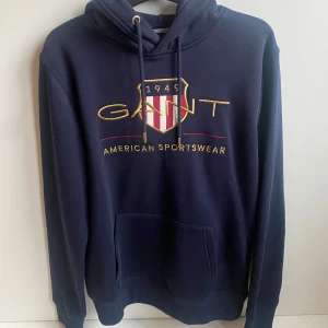 Mörkblå hoodie från GANT - Säljer en snygg mörkblå Gant Shield hoodie med stort tryck på framsidan. Den har en klassisk känguruficka och justerbar huva med snören. Perfekt för en avslappnad stil eller kyliga dagar. Passar både till vardags och sportiga aktiviteter. Inköpt för 1300kr