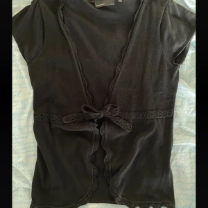 Svart kortärmad topp från KappAhl  - Snygg svart kortärmad topp från KappAhl med knytband i midjan. Perfekt för både vardag och fest. Den har en fin spetsdetalj längs kanterna som ger en extra touch. Storlek M