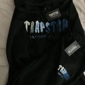 Säljer en svart hoodie från Trapstar med coolt tryck i blått och vitt på framsidan. Den har en klassisk passform med huva och dragsko. Perfekt för en avslappnad stil eller streetwear-look. Helt ny med taggar kvar!