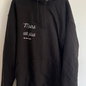 Svart hoodie med tryck  - Säljer en svart hoodie med texten 'Moins est plus' tryckt på både fram- och baksidan. Den är i bra skick och perfekt för en avslappnad stil. Hoodien har en klassisk passform med långa ärmar och en bekväm huva. Perfekt för höst och vinter!