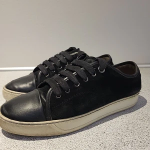 Svarta sneakers från Lanvin strl 41 - Säljer ett par svarta sneakers från Lanvin i skinn och mocka. De har en klassisk design med snörning och en vit sula som ger en snygg kontrast. Perfekta för både vardag och fest! Lite använda men i bra Innermått 26 cm