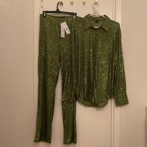 Grön paljettbyxa och skjorta från Gina Tricot - Säljer ett set med gröna paljettbyxor och matchande skjorta från Gina Tricot. Perfekt för festliga tillfällen! Byxorna har en bekväm passform och skjortan har knappar framtill. Båda plaggen är i nyskick och redo att glittra på nästa fest. Stl L