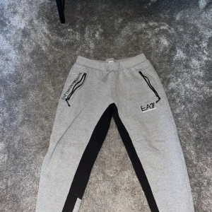 Grå mjukisbyxor från Emporio Armani - Säljer ett par grå mjukisbyxor från Emporio Armani EA7. De har svarta detaljer längs benen och dragkedjor vid fickorna. Perfekta för en avslappnad stil eller träning. Byxorna har en elastisk midja med snörning för bästa passform.
