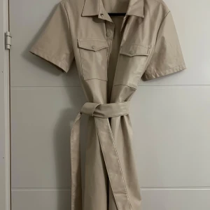 Klänning i läderimitation  - Klänning i läderimitation som är i väldigt fint skick, inga anmärkningar alls. Beige, knytning, knappar framtill och fickor vid brösten. Jag är 167 cm och den slutar några cm ovanför knäna på mig 