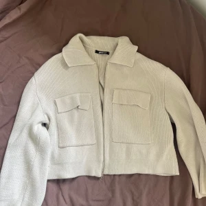 Beige stickad jacka med dragkedja - Säljer en superfin beige stickad jacka med dragkedja framtill. Den har en croppad passform och två stora fickor på bröstet. Perfekt för höst och vår när du vill ha något stilrent och bekvämt. Jackan är långärmad och har en klassisk krage.