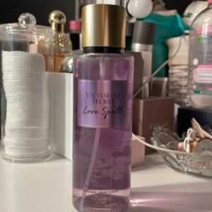 Victoria's Secret Love Spell Body Mist - Säljer en fräsch och förförisk body mist från Victoria's Secret i doften Love Spell. Den har en ljuvlig blandning av körsbärsblom och färsk persika som är omöjlig att motstå. Flaskan är genomskinlig med en lila ton och har en elegant guldlock. Perfekt för daglig användning eller speciella tillfällen!