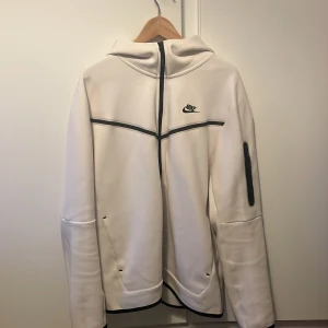 Vit hoodie från Nike - Säljer en snygg vit hoodie från Nike med dragkedja och huva. Den har svarta detaljer och en ficka på ärmen. Perfekt för både träning och vardag!
