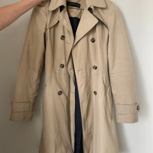Trenchcoat - Säljer en klassisk beige trenchcoat från Zara i storlek S. Den är i fint skick och i stilren design med knappar och ett bälte i midjan. Perfekt för vår och höst!