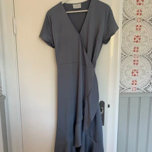 NeoNoir omlottklänning - NeoNoir ”Magga dress”, gråblå omlottklänning med volang. Använd två gånger så fint skick. Ett litet märke på ärmen som knappt syns (vg se bild 3). 