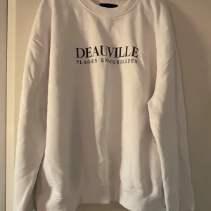 Vit sweatshirt från 157 - Säljer en stilren vit sweatshirt från lager157 med texten 'DEAUVILLE PLAGES ENSOLEILLÉES' tryckt på framsidan. Tröjan har en normal passform och är perfekt för både vardag och avslappnade tillfällen. Den är i bra skick och superbekväm! Hör gärna av er vid frågor eller fler bilder💕